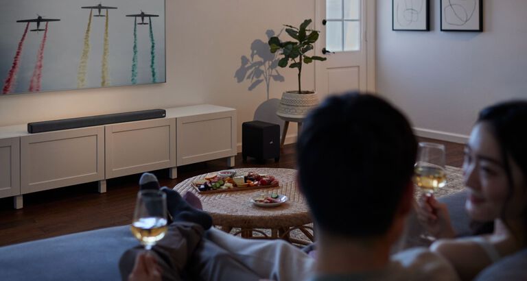 a couple watching TV with a large sound bar sitting under it.